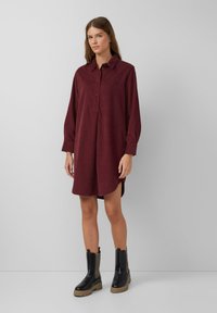 Robe chemise bordeaux à manches longues, avec un devant à boutons et des fentes sur les côtés, associée à des bottines noires avec des panneaux texturés.