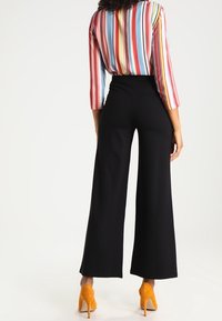 Szerokie czarne spodnie z gładkiego materiału, zestawione z wielokolorową, paskowaną bluzką z rękawami do łokcia oraz żółtymi butami na obcasie.