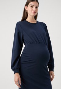 Rochie de maternitate bleu marin cu mâneci lungi, talie strânsă și țesătură moale. Dispune de un decolteu rotund și buzunare laterale pentru o funcționalitate suplimentară.