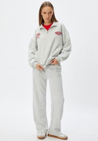 Sudadera gris con cuello de media cremallera, detalles bordados y dos bolsillos frontales, combinada con pantalones deportivos grises a juego y zapatillas blancas.