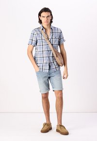 Korte mouwen geruit shirt in blauw en wit, lichte denim shorts en bruine suède schoenen. Model staat met een beige crossbody tas.