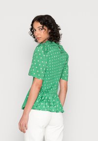 Blusa peplum verde com mangas curtas, apresentando um costado franzido, padrões florais brancos e um fecho em forma de olho-chave na gola.
