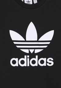 Camiseta de algodón negra con un gran logo blanco de Adidas que presenta tres franjas similares a hojas sobre el nombre de la marca. Diseño clásico de cuello redondo.