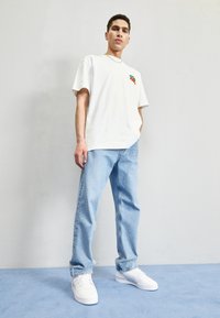 Junger Mann steht an einer Wand und trägt ein weißes T-Shirt mit Kirschdesign, hellblaue Jeans, weiße Sneaker und eine dünne Halskette.