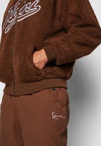 Brown fleece zip-up hoodie with ribbed cuffs and hem; features a white logo. Paired with brown pants, showcasing a white embroidered detail.