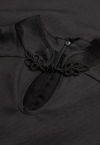 Schwarzes Kleidungsstück mit strukturiertem Stoff, hohem Kragen, Reißverschluss und dekorativen Knotenakzenten in der Nähe der Öffnung.