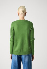 Grüner gestrickter Pullover mit rundem Halsausschnitt und geripptem Saum, ausgestattet mit langen Ärmeln und lockerer Passform. Getragen mit blauen weiten Jeans.