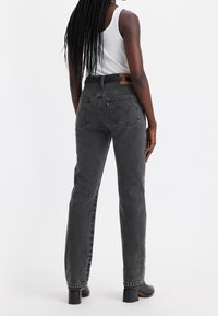 Jeans de tiro alto en gris oscuro con piernas rectas, bolsillos traseros y una etiqueta de cuero en la cintura, combinados con zapatos negros de tacón.