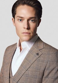 Brązowa marynarka w kratę o teksturowanym wykończeniu, z klapami w kształcie notcha i czarnym detalem na lewej stronie. Pod spodem biała koszula.