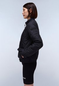 Black quilted jacket with a high collar, side pockets, and a fitted design. Worn over black cycling shorts, showcasing a smooth texture.