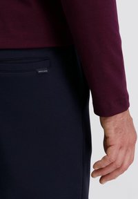 Bordeaux langslieve shirt gecombineerd met marineblauwe broeken. Close-up van de linkerarm en hand, met zicht op de mouw en een zaklabel.