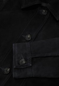 Manche et devant d'une veste en daim noir avec boutons noirs et détails de couture visibles.