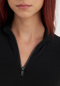 Crna zip-up majica s visokim ovratnikom, s teksturiranim zatvaračem. Tkanina izgleda glatko i priljubljeno, naglašavajući liniju vrata.
