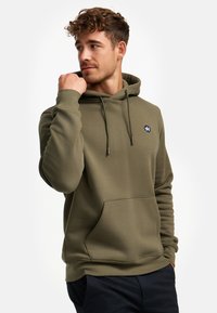 Junger Mann mit lockigem Haar, der einen olivgrünen Kapuzenpullover trägt, eine Krempe nahe seinem Kinn hält und zur Seite blickt, vor einem weißen Hintergrund.