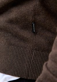 Brun strikket genser med ribbet kant og liten svart rektangulær merkelapp merket "JACK & JONES" på stoffet nær bunnen.