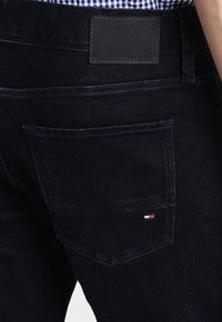 Dunkelblaue Jeans mit glatter Textur, ausgestattet mit einem Leder-Logopatch am Bund und einer Gesäßtasche mit einem kleinen Markenlogo-Tag.