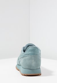 Ljusblå mockasko med en texturerad häl och vadderad krage. Har en tan gummisula och präglar Reebok-logotypen på baksidan.