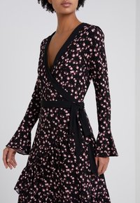 Musta kukallinen kietaisumekko, jossa on vaaleanpunaisia ruusukuviota, levenevät hihat ja solmittava vyötäröyksityiskohta. Mekossa on röyhelöhelma ja pehmeä kangas.