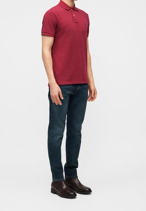 Mann trägt ein rotes Polohemd, dunkelblaue Jeans und schwarze Lederstiefel, steht vor einem weißen einfarbigen Hintergrund.
