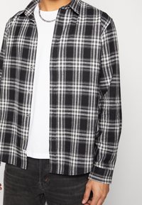 Sort og hvid ternet flannel skjorte med knap-ned design og lange ærmer. Båret over en ensfarvet hvid t-shirt og parret med mørke jeans.