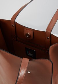 Brązowa torba na ramię z imitacji skóry, z dwoma uchwytami, zapięciem na zatrzask oraz kieszeniami wewnętrznymi. Posiada tłoczoną teksturę i zawieszkę z logo wewnątrz.