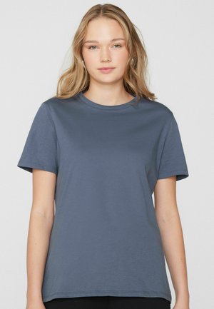 Mladá žena s dlouhými blond vlasy, oblečená v jednoduchém modrém tričku s krátkým rukávem, na světlém pozadí.