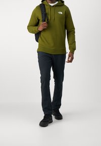 Olijfgroene hoodie met trekkoord, logo op de borst, gecombineerd met donkere broeken en zwarte robuuste schoenen. Rugzak gedragen op de linkerschouder.