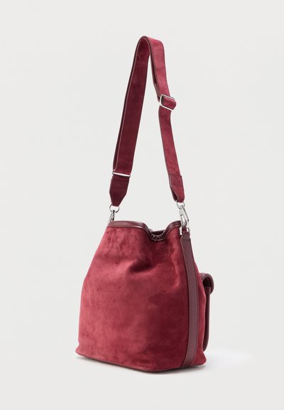 Κόκκινη τσάντα τύπου πουγκί από σουέτ με ρυθμιζόμενο ιμάντα ώμου και ασημένια μεταλλικά στοιχεία, παρουσιάζεται σε απλό φωτεινό φόντο.