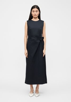 Femme debout portant une robe midi noire sans manches avec un détail de nouage sur le côté et des escarpins blancs à bout pointu sur un fond clair uni.