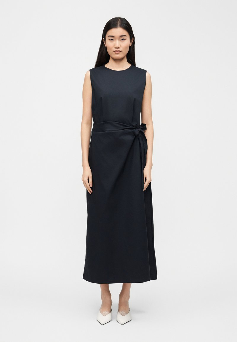 Femme debout portant une robe midi noire sans manches avec un détail de nouage sur le côté et des escarpins blancs à bout pointu sur un fond clair uni.