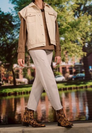 Beige denim vest with large pockets, brown sweater underneath, white distressed jeans, and brown snake-pattern ankle boots.