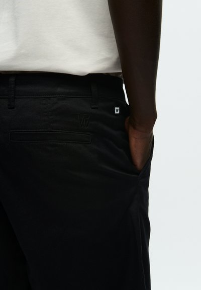 Pantalon noir avec une texture lisse, une coupe décontractée, des poches latérales et un logo subtil sur la poche arrière. Tissu en mélange de coton.