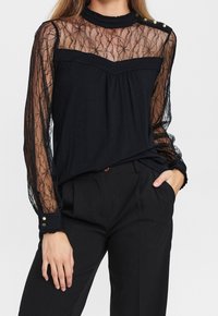 Femme portant un haut en dentelle noire transparente à manches longues avec des détails en boutons dorés, associé à un pantalon noir ajusté, les mains dans les poches.