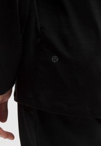 Detailný záber na ruku osoby a čierne športové oblečenie, zobrazené s malým reflexným logom Lululemon v blízkosti lemu.