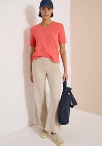 Femme portant un t-shirt corail à manches courtes, un pantalon beige à jambes larges, des baskets crème et une casquette bleu marine, tenant un sac à dos bleu marine à ses côtés.