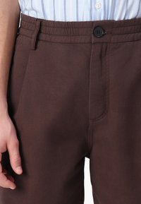 Braune Baumwollhose mit elastischem Bund, ausgestattet mit einer Seitentasche und einem schwarzen Knopfverschluss. Glatte Textur, gerade geschnittenes Design.
