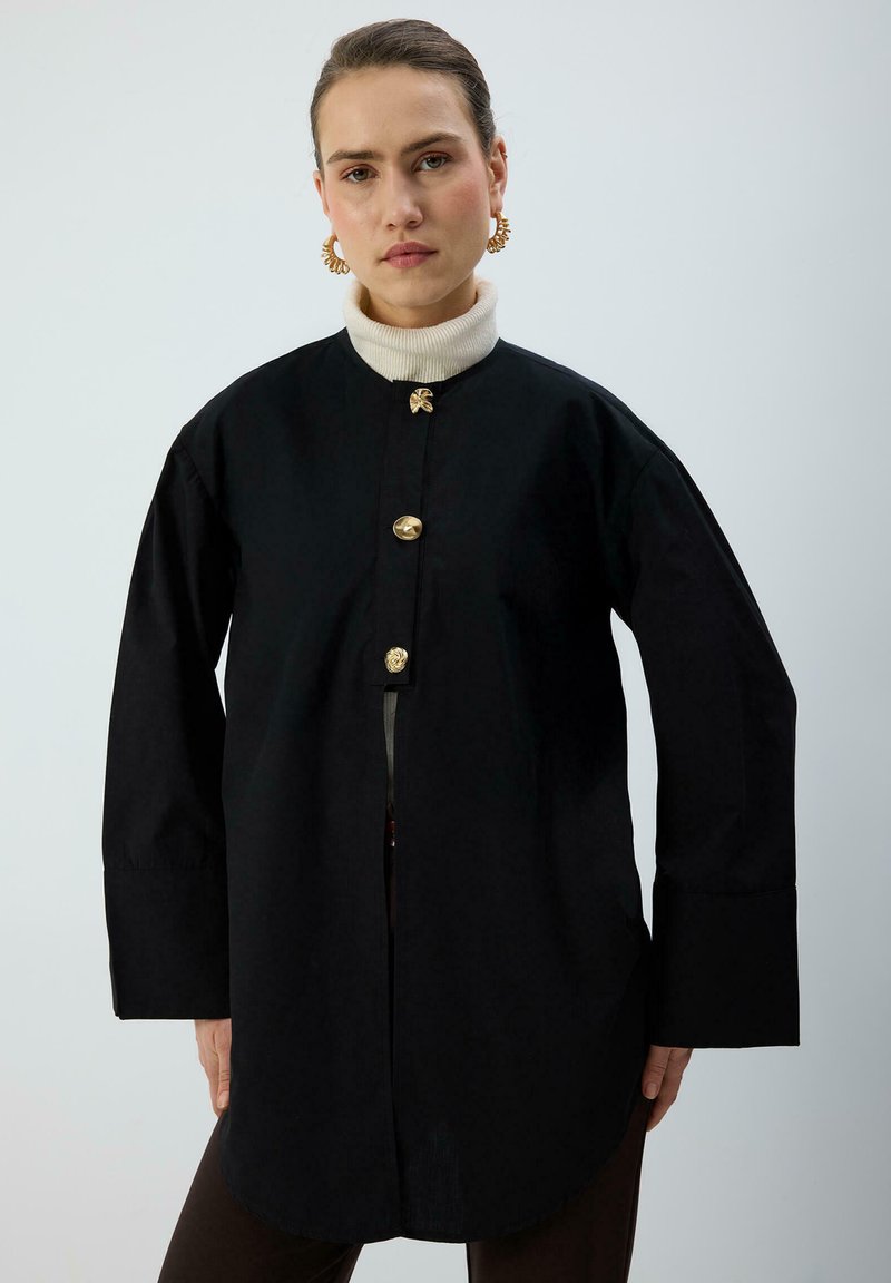 Camicia di cotone nera con bottoni dorati, colletto alto e maniche lunghe. Presenta un orlo dritto e una vestibilità morbida. Nessun motivo distintivo.