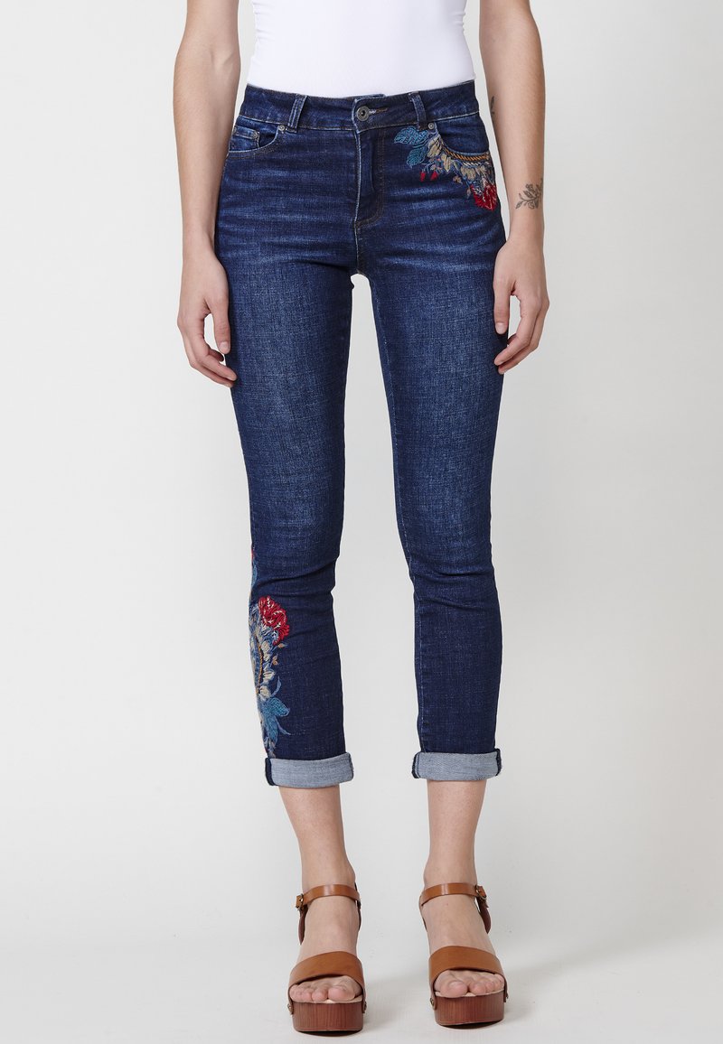 Jeans de mezclilla de lavado oscuro con puños doblados, que presentan bordados florales coloridos en el muslo izquierdo. Diseño ajustado con estilo de cinco bolsillos.