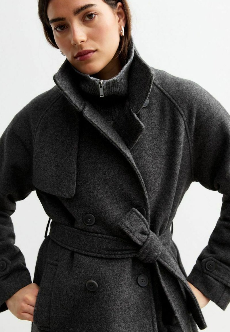 Cappotto in misto lana grigio scuro con design doppiopetto, grandi bottoni e vita con cintura. Presenta un colletto alto e un lavoro a maglia testurizzato sotto.