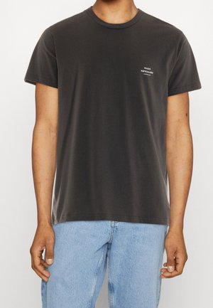 Homme portant un t-shirt à col rond gris foncé avec un petit texte blanc sur la poitrine gauche et un jean bleu clair, se tenant avec les bras détendus.