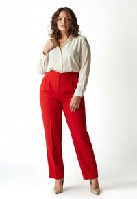 Weiße Bluse mit langen Ärmeln und Knöpfen, kombiniert mit hochtaillierter, plissee-roter Hose. Die Schuhe sind metallisch beige. Schlichter Hintergrund.