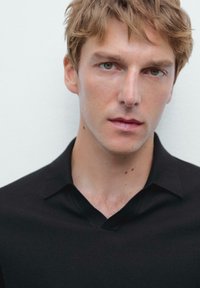 Černá pletená polo košile s véčkovým výstřihem, minimalistickým designem a krátkými rukávy. Model má světle hnědé vlasy a pleť neutrální barvy s pihami.