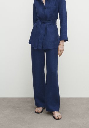 Femme portant une veste bleu marine ceinturée avec un pantalon large assorti et des sandales noires à brides, debout sur un sol gris contre un mur blanc.