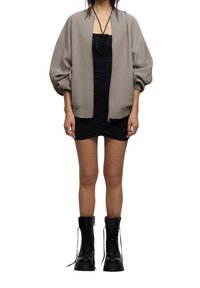 Chaqueta bomber gris sobre un vestido mini negro con fruncidos, combinada con botas de tobillo negras con cordones. La chaqueta tiene un cierre de cremallera en la parte delantera y mangas relajadas.