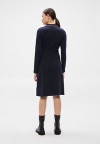 Tumši zila kleita ar garām piedurknēm, vidukļa iespaidu, apkakles detaļu un taisnu siluetu, kombinēta ar melniem potīšu zābakiem.