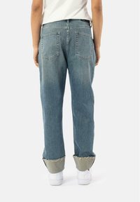 Denim jeans with a relaxed fit, light blue color, and frayed hem. Features two back pockets and worn detailing at the cuffs, paired with white shoes.