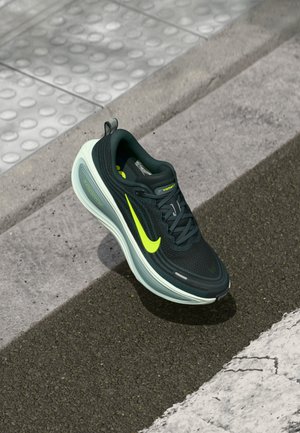 Pantofii de alergare Nike, negri cu verde fluorescent, dotati cu talpa ZoomX, plutind aproape de un bordură și o suprafață cu pavele tactile.
