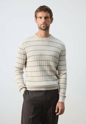 Hombre con cabello corto y castaño y barba, que lleva un suéter de punto con ochos color crema con rayas horizontales y pantalones oscuros, de pie con una mano en el bolsillo.