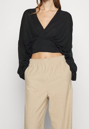 Vrouw met een zwart, gerimpeld crop top met lange mouwen en een V-hals, en beige broek met elastische taille, staand met handen in de zakken.