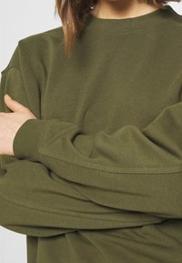 Sudadera de gran tamaño en verde oliva, hecha de un tejido suave, con cuello redondo y costuras de hombro caídas, que presenta puños y dobladillo de canalé.